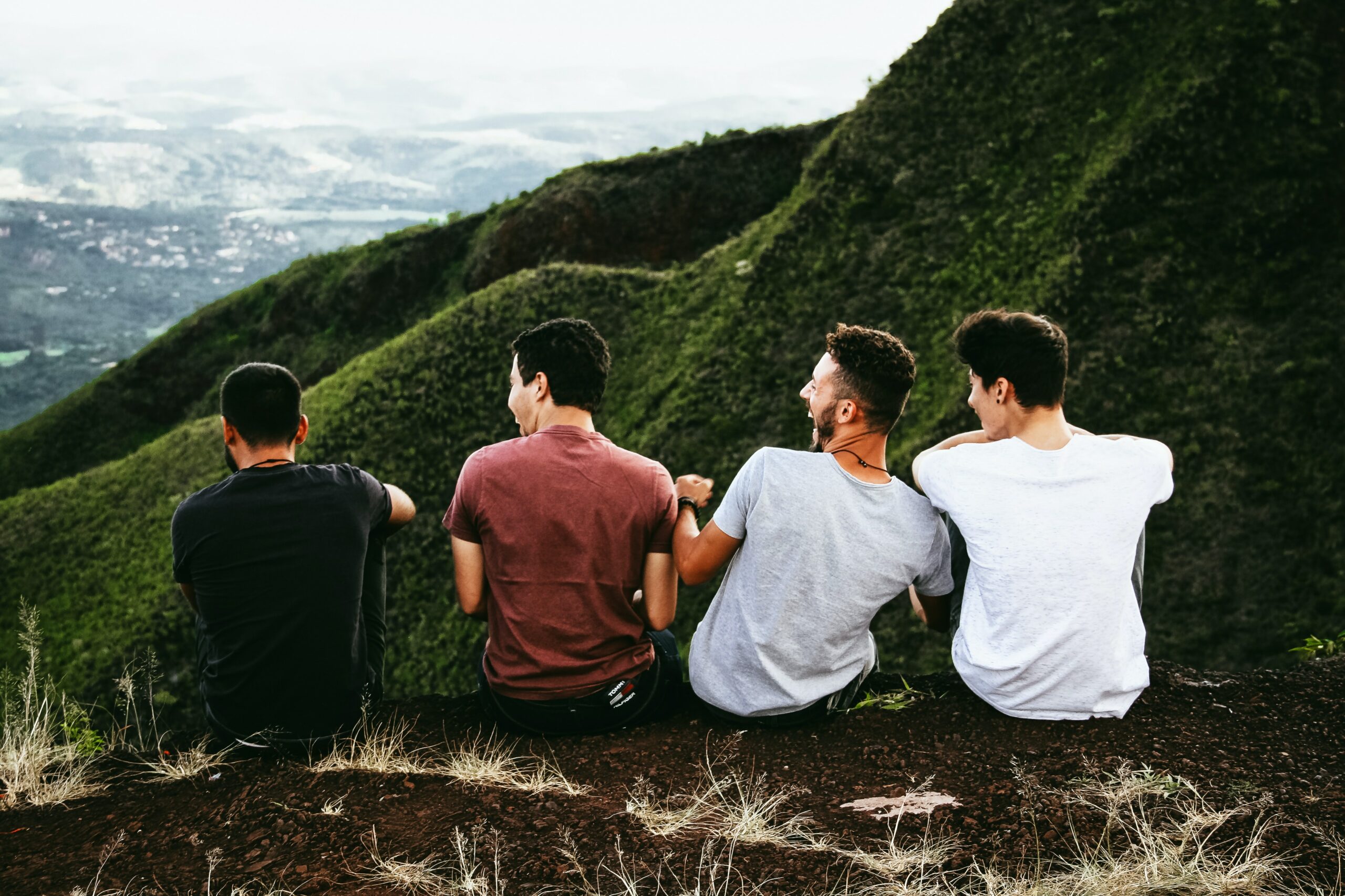 Vier junge Männer sitzen dem Betrachter mit dem Rücken zugewandt nebeneinander in der Natur und lachen